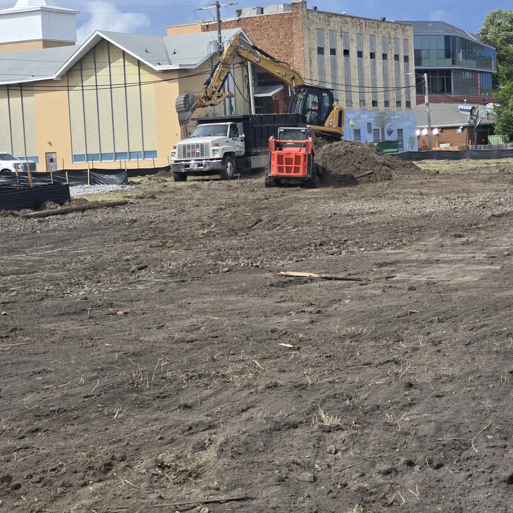 Excavator, dump truck and compact track loader working within a fenced urban construction site.