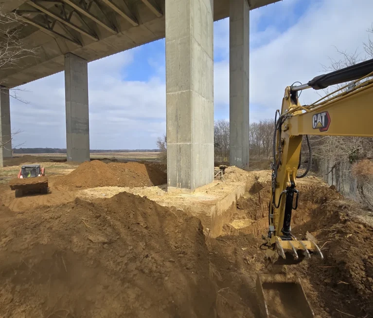 Excavator and tracked loader performing earthwork beneath highway bridge spans.