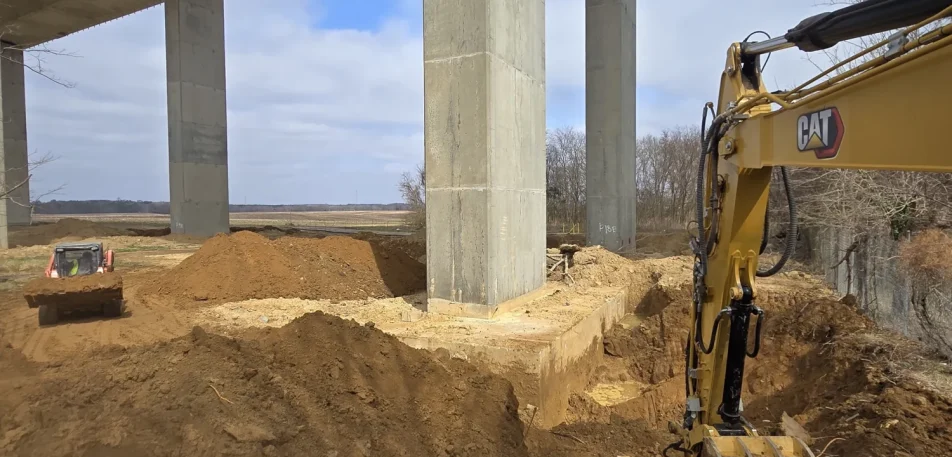 Excavator and tracked loader performing earthwork beneath highway bridge spans.