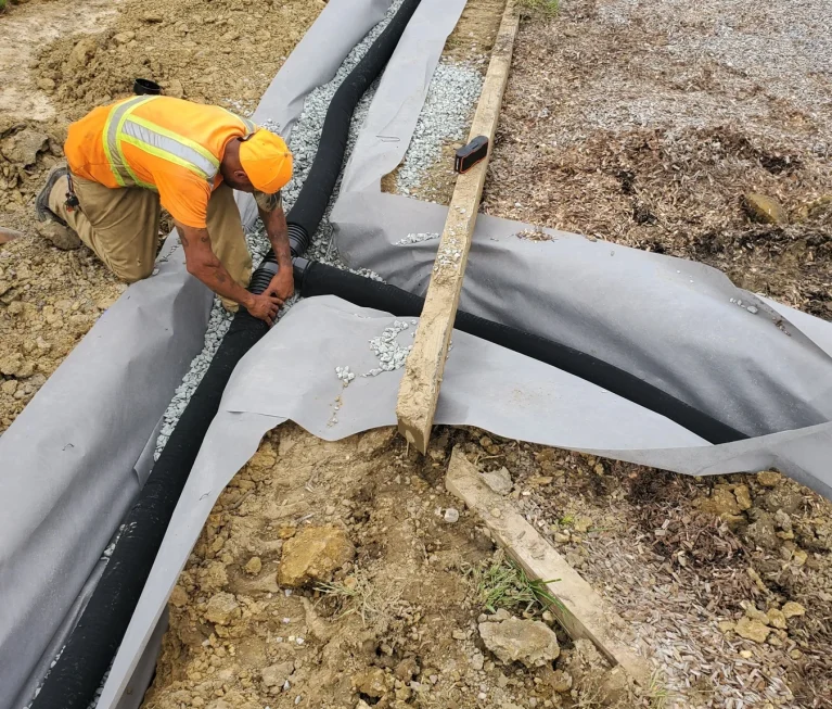 Worker joining corrugated pipe in a fabric-lined gravel trench.
