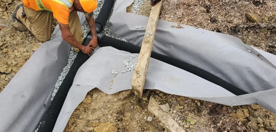 Worker joining corrugated pipe in a fabric-lined gravel trench.