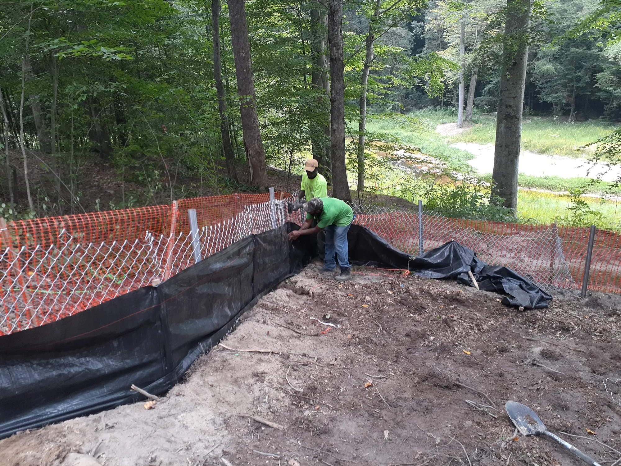 Two crew members installing black silt-fence fabric beside a safety barrier.